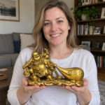 Woman in her thirties supporting the gold-finished copper Laughing Buddha with money bag in both hands at home